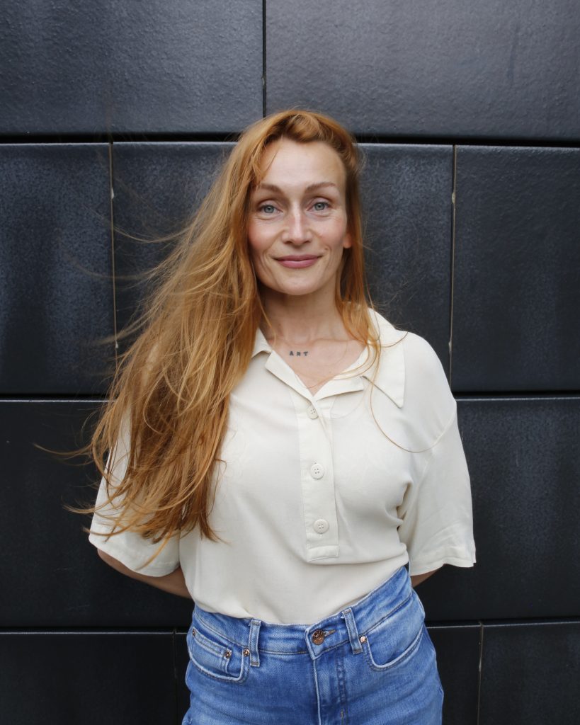 Portrait Sarah Haack, Fliesen als Hintergrund, sie trägt eine helle Bluse und Jeans.