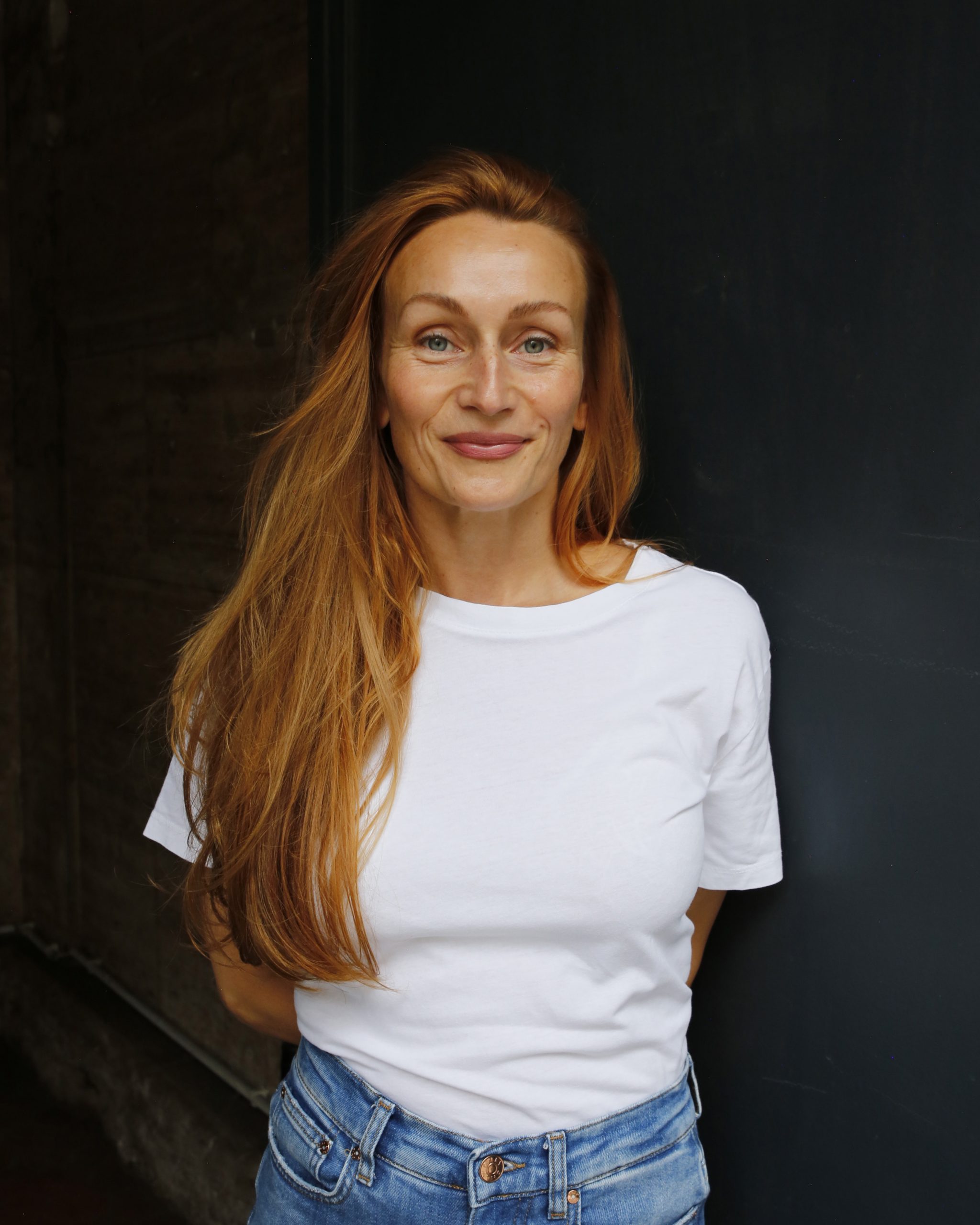 Portrait Sarah Haack, dunkler Hintergrund, sie trägt ein weißes T-Shirt und Jeans.