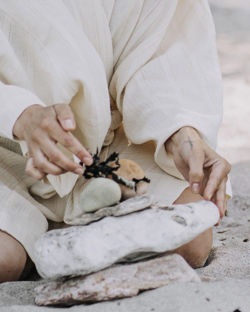 Nahaufnahme, Person kniet im Sand und stapelt Steine. Sie trägt ein helles locker sitzendes Gewand.